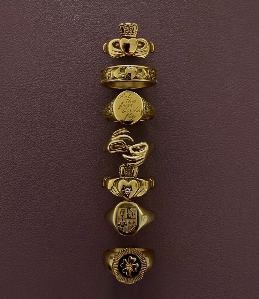 A vertical stack of seven Irish gold signet rings with various heraldic and decorative engravings, displayed on a textured dark plum background.